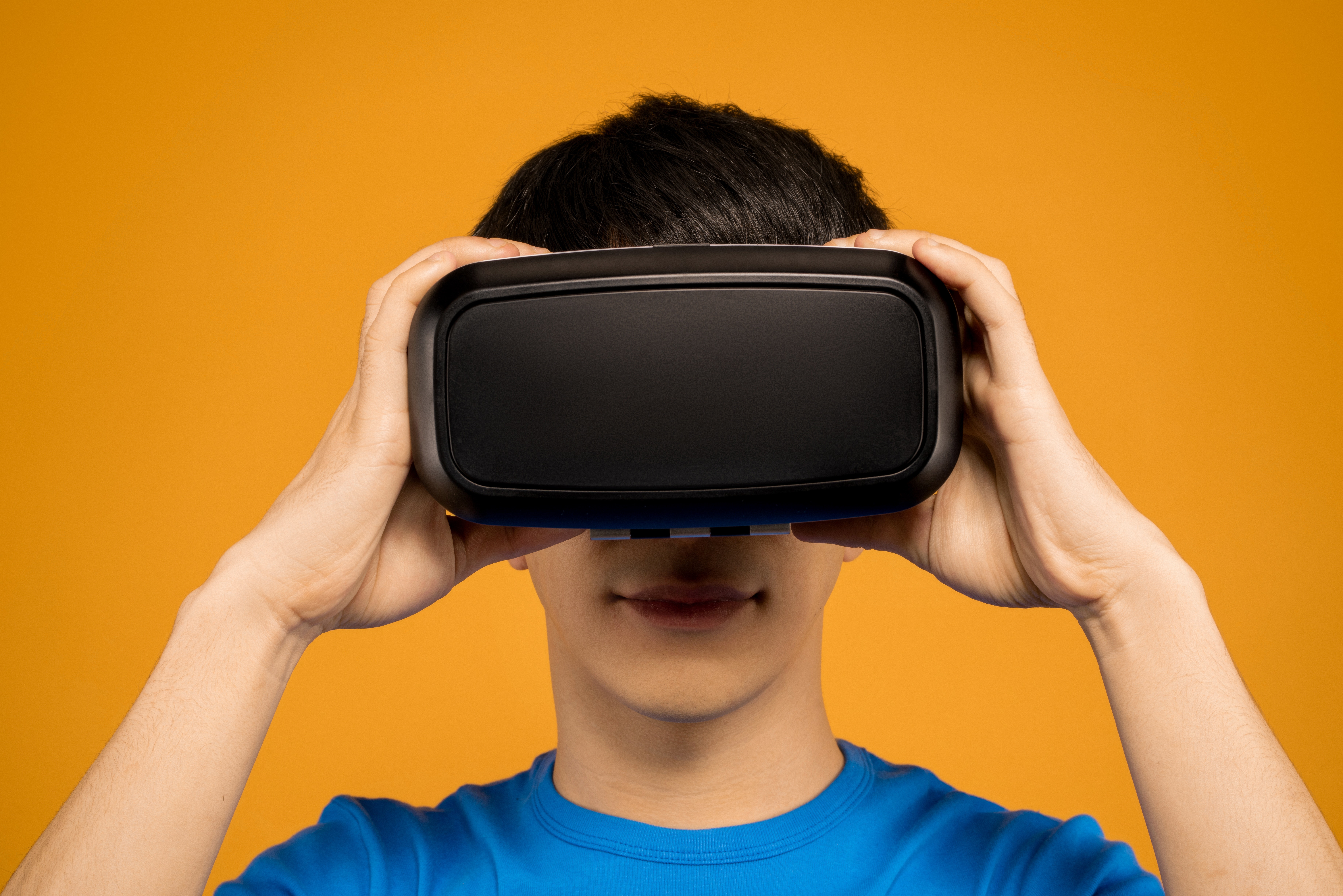 Image shows a person in a blue t-shirt holding a VR headset up to their face against a yellow background.